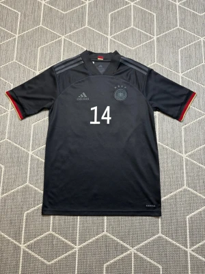 Tyskland Adidas - Snygg svart matchtröja från Adidas med Tysklands landslagsmärke och nummer 14 på bröstet. Tröjan har korta ärmar, gråa ränder på axlarna och detaljer i rött och gult vid ärmsluten. Tillverkad i lätt och ventilerande material, perfekt för fotboll.