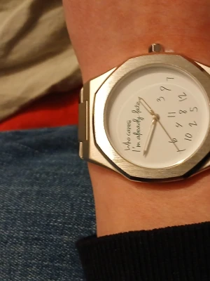 Rolig silverfärgad klocka - Unik klocka i silverfärgad metall med vitt ur och texten 'Who cares I'm already late'. Siffrorna är lekfullt placerade i en hög på urtavlan. Klockan har en klassisk länkarmband och modern, kantig form. Perfekt för dig som gillar humoristiska accessoarer.