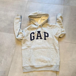 Grå hoodie från GAP - Säljer en klassisk grå hoodie från GAP med texten 'GAP ITALY' i svart och vitt på bröstet. Tröjan har en stor ficka framtill och justerbar huva med snören. Perfekt för en avslappnad stil. Bara att höra av sig vid funderingar!🤩