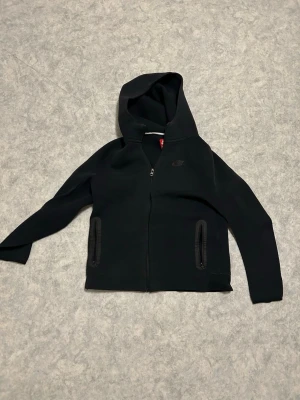 Svart hoodie med dragkedja från Nike - Svart hoodie från Nike med dragkedja framtill och två fickor med dragkedja. Klassisk design med huva och diskret Nike-logga på bröstet. Perfekt för en sportig och avslappnad stil. Tillverkad i mjukt och bekvämt material.