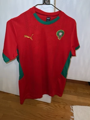 Marocko fotbollströja Puma röd/grön - Säljer en officiell Marocko fotbollströja från Puma i rött med gröna detaljer. Tröjan har korta ärmar, broderad Puma-logga och Marockos landslagsemblem på bröstet. Tillverkad i lätt och ventilerande polyester, perfekt för träning eller match. Passar som S 