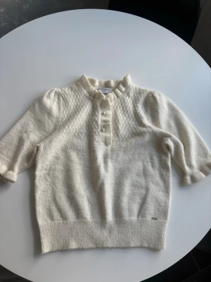 Vit stickad tröja Holly & Whyte - Söt vit stickad tröja från Holly & Whyte med korta puffärmar och ribbad kant. Tröjan har ett diskret mönster upptill och pärlknappar vid halsen. Perfekt till både jeans och kjol för en stilren look.