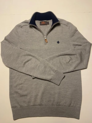 Ljusgrå halvzip-tröja från Morris - Säljer en stilren ljusgrå stickad tröja från Morris i 100% merinoull. Tröjan har halv dragkedja, mörkblå krage och en liten broderad logga på bröstet. Perfekt för dig som gillar klassisk och bekväm stil.