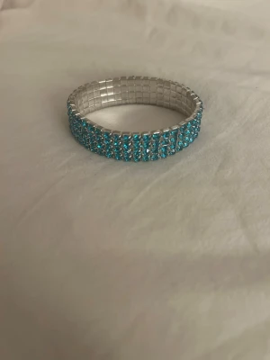 Elastiskt armband med blå pärlor - Snyggt elastiskt armband med flera rader små, glittrande blå pärlor på en silverfärgad bas. Perfekt för att ge din outfit lite extra färg och bling. Armbandet har en flexibel design som gör det enkelt att ta på och av.