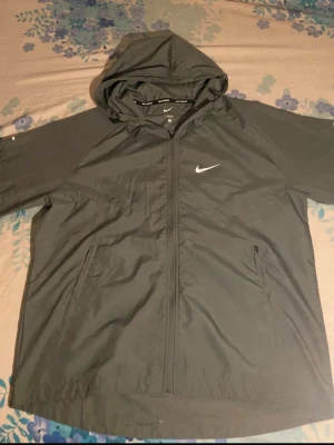 Grå nike Miller windbraker - Svart vindjacka från Nike med dragkedja framtill, två sidofickor och justerbar huva. Jackan har en stilren design med Nike-logga på bröstet och är tillverkad i lätt, vindtätt material. Perfekt för blåsiga dagar och sportiga outfits.