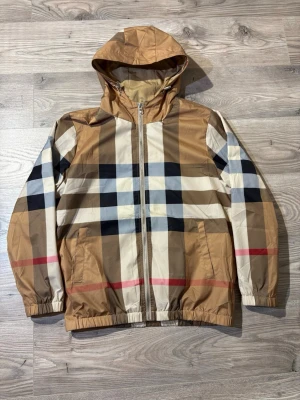 Beige rutig vindjacka från Burberry - Snygg beige vindjacka från Burberry med klassiskt rutmönster i svart, vitt, rött och brunt. Jackan har huva, dragkedja framtill och två fickor. Tillverkad i lätt och tåligt material, perfekt för blåsiga dagar. Elastiska muddar vid ärmslut och nederkant. XL junior, passar herr S/M