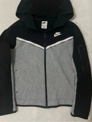 Nike Tech - Nike hoodie i svart och grå med vit logga på bröstet och hel dragkedja framtill. Huva och långa ärmar, samt vit detalj över bröstet. Tillverkad i mjukt material, perfekt för en sportig och avslappnad stil.