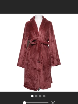 Vinröd mjuk morgonrock med skärp - Mysig vinröd morgonrock i fluffigt, sammetsliknande material. Klassisk modell med sjalkrage, långa ärmar och knytskärp i midjan. Perfekt för att chilla hemma och hålla sig varm under kyliga morgnar. Köpt på hunkemöller!!