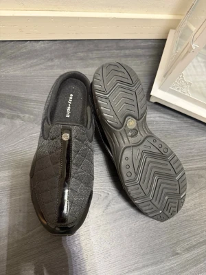 Svarta quiltade tofflor Easy Spirit - Säljer ett par svarta tofflor från Easy Spirit med quiltat mönster och en blank detalj framtill. Tofflorna har en mjuk innersula och rejäl gummisula för extra komfort. Perfekta för att glida runt hemma i stil.