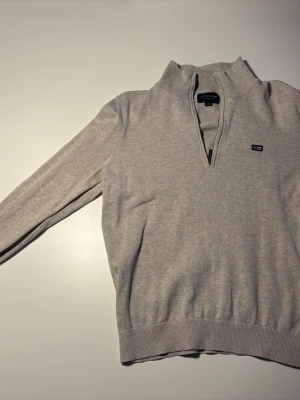 Ljusgrå halvzip-tröja Lexington - Ljusgrå stickad tröja från Gant med halvzip och hög krage. Tröjan har ribbade muddar vid ärmslut och nederkant samt en liten Gant-logga på bröstet. Perfekt för lager på lager och enkel att matcha med olika outfits.