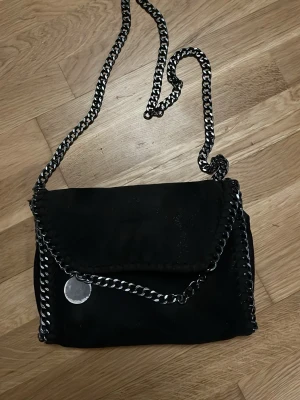 Svart axelväska med kedjedetaljer - Snygg svart axelväska med silverfärgad kedja som axelrem och runt kanterna. Väskan har ett lock med magnetknäppning dock har ena delen lossnat så den kan inte stängas. Materialet ser ut som mockaimitation och ger en cool vibe till outfiten.