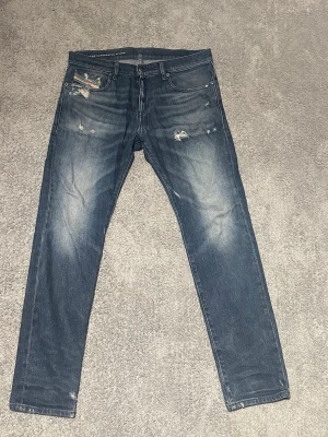 Blå slitna jeans från Diesel - Säljer ett par blå jeans från Diesel med slitningar och distressed detaljer på fram- och baksidan. Jeansen har klassisk femficksdesign och smal passform. Perfekta för en avslappnad och trendig look. Storlek w31 L30