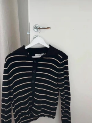 Svart stickad tröja med ränder - Snygg svart stickad tröja från NA-KD med vita horisontella ränder och knappar framtill. Tröjan har krage och långa ärmar, perfekt för en trendig och avslappnad look. Endast använd en gång!!