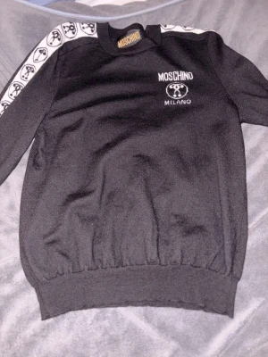 Svart Moschino sweatshirt med logga - Snygg svart sweatshirt från Moschino med vit logga och Milano-tryck på bröstet. Tröjan har ribbade muddar och vita band med Moschino-mönster längs axlarna. Perfekt för dig som gillar streetwear och exklusiva märken.