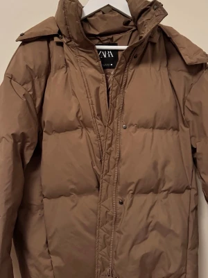 Brun pufferjacka från ZARA - Säljer en brun pufferjacka från ZARA med hög krage och dragkedja framtill. Jackan har en klassisk pufferdesign med breda kanaler och tryckknappar. Perfekt för kalla dagar och riktigt skön att bära. Materialet är glansigt och vattenavvisande.