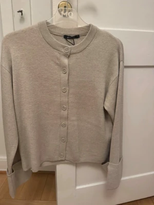 Beige melerad kofta med knappar - Stilren beige melerad kofta med rund hals och knäppning framtill. Tröjan har långa ärmar och ribbade muddar vid ärmslut och nederkant. 