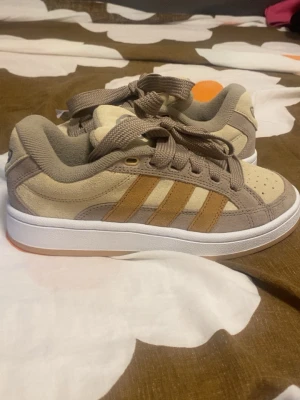 Beige Adidas sneakers med mockadetaljer - Snygga beige sneakers från Adidas med klassiska tre ränder i brunt på sidan. Skorna har ovandel i mocka och textil, samt vita sulor och beige snörning. Perfekta för dig som gillar en clean och stilren look.