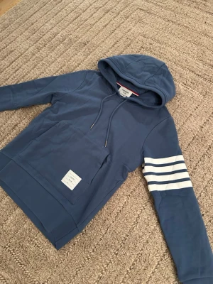 Blå hoodie från Thom Browne - Snygg blå hoodie från Thom Browne med klassiska vita ränder på ena ärmen och en patch framtill. Hoodien har dragsko i huvan och en stor magficka. Tillverkad i mjuk bomull för skön känsla och avslappnad stil.