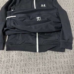 Svart loungewear-set från Under Armour - Säljer ett svart loungewear-set från Under Armour med dragkedja på tröjan och matchande byxor. Setet har vita detaljer och logga på både tröja och byxa. Perfekt för chill dagar hemma eller när du vill vara bekväm men ändå se snygg ut.