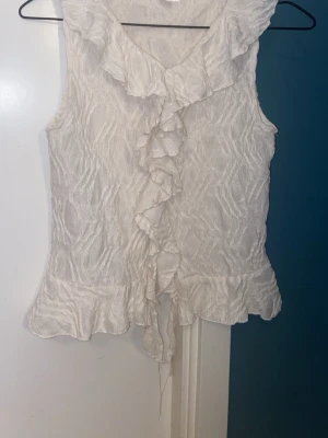 Vit ärmlös blus med volang - Supersöt vit blus i ett strukturerat tyg med volangdetaljer längs kragen och framtill. Ärmlös modell med v-ringning och volangkant nedtill. Är i storlek 170 men skulle säga att den passar xs också! Pris kan diskuteras 💕