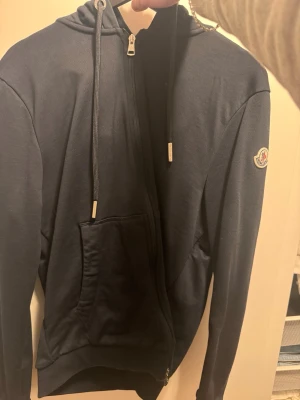 Svart hoodie med dragkedja från Moncler - Snygg svart hoodie från Moncler med dragkedja framtill, huva med dragsko och klassisk logotyp på ärmen. Hoodien har en stor magficka och ribbade muddar. Perfekt för en avslappnad och trendig stil.