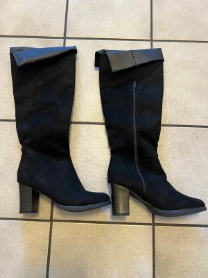 Svarta höga stövlar med klack  - Snygga svarta stövlar med bred klack och uppvikt kant upptill. Dragkedja på insidan för enkel påtagning. Materialet ser ut att vara mockaimitation med detaljer i konstläder. Perfekta för att lyfta din outfit med en edgy touch.