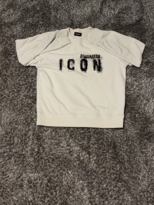 Vit Dsquared2 ICON hoodie - Vit hoodie från Dsquared2 med svart text 'ICON' och Dsquared2-logga på bröstet. Tröjan har klassisk passform, ribbade muddar och rund halsringning. Materialet är mjukt och bekvämt, perfekt för en avslappnad streetwear-look.