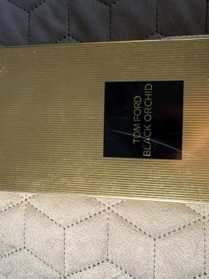 Tom Ford Black Orchid parfym - Lyxig parfym från Tom Ford, Black Orchid, i en elegant guldfärgad förpackning med svart etikett. Förpackningen har en stilren, rektangulär form och är tillverkad av kartong med räfflad yta. Perfekt för dig som gillar exklusiva dofter och snygg design.