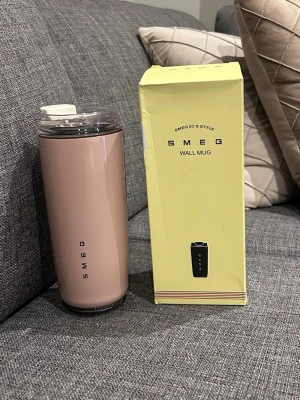 Smeg rosa termosmugg med lock - Snygg rosa termosmugg från Smeg med lock, perfekt för att ta med kaffe eller te på språng. Muggen har en stilren design med Smeg-logga och kommer i originalförpackning. Tillverkad i rostfritt stål och plast, rymmer ca 500 ml.