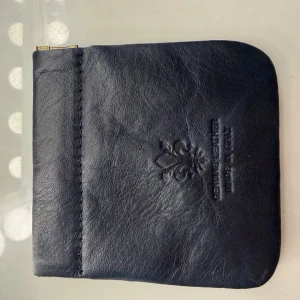 Svart läderplånbok med präglad detalj - En stilren svart plånbok i äkta läder med präglad dekor på framsidan. Plånboken har en enkel, fyrkantig form och dragkedja upptill. Märkt med 'Genuine Leather, Made in Italy'. Perfekt för kort och mynt.