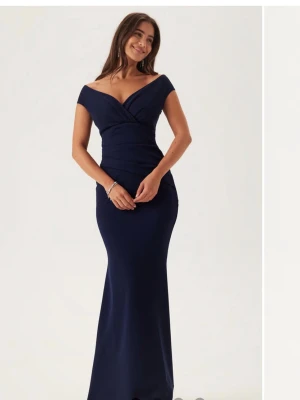 Marinblå långklänning offshoulder - Elegant marinblå långklänning med offshoulder och v-ringad omlott över bysten. Den är aldrig använd och har kvar lappen. Klänningen har figurnära passform och är tillverkad i ett mjukt, stretchigt material som ger en snygg siluett. Perfekt för speciella tillfällen som tex bal.