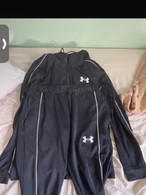 Svart träningsset från Under Armour - Säljer ett svart träningsset från Under Armour med vita detaljer och logga på både jacka och byxor. Setet har dragkedja på jackan, muddar vid ärmslut och elastisk midja på byxorna. Perfekt för gymmet eller löprundan.