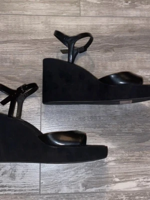 Svarta kilklackssandaler från H&M - Säljer ett par svarta kilklackar från H&M, med justerbar rem runt ankeln. Ovandelen är i slätt konstläder och sulan är i ett mattare sammet material. Snygg minimalistisk design med öppen tå och bred rem framtill.