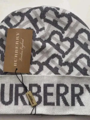 Gråvit Burberry mössa med logomönster - Säljer en snygg gråvit mössa från Burberry med stort logomönster och tydlig Burberry-text framtill. Mössan är stickad och har en modern, avslappnad passform. Perfekt accessoar för att lyfta din outfit under kyliga dagar.