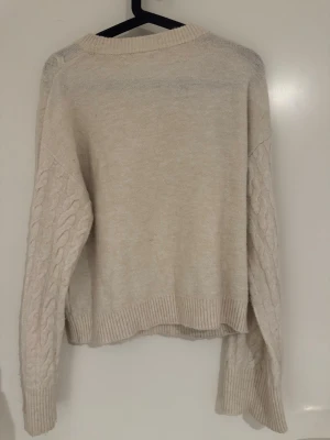 Beige stickad tröja från H&M XS - Beige stickad tröja från H&M i storlek XS. Tröjan har flätstickade ärmar och ribbade muddar vid ärmslut och nederkant. Perfekt för lager-på-lager och har en avslappnad passform. Materialet är mjukt och bekvämt.