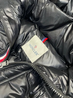 Svart Moncler pufferjacka med dragkedja - Säljer en svart pufferjacka från Moncler med glansig finish och breda, quiltade sektioner. Jackan har en kraftig dragkedja, coolt innerfoder med serietidningsmotiv och Moncler-logga. Röd, vit och marinblå detalj vid dragkedjan. Perfekt för kalla dagar.