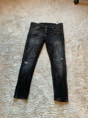 Svarta slitna jeans från Dsquared2 - Säljer ett par svarta jeans från Dsquared2 med slitningar och tvättade detaljer. Jeansen har smal passform, klassisk femficksmodell och är tillverkade i denim. Perfekta för en trendig och avslappnad stil.