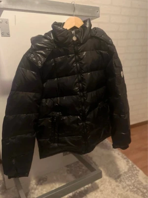 Svart dunjacka med nytt skickt  - Snygg svart dunjacka med hög krage och dragkedja framtill. Jackan har två framfickor med dragkedja och quiltad design. Passar perfekt för kalla vinterdagar och har en modern, puffig look. Märke syns på ärmen.