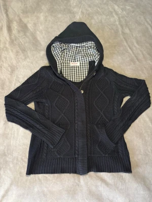 Svart stickad hoodie - Svart stickad hoodie med dragkedja och ribbade muddar. Snyggt rutigt innerfoder i huvan och klassiskt stickat mönster med stora rutor. Perfekt för dig som gillar en avslappnad och mysig stil.