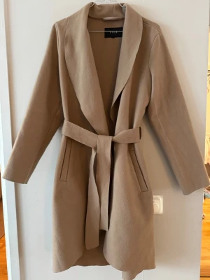 Beige kappa med skärp från Vila - Stilren beige kappa från Vila med knytskärp i midjan och två fickor framtill. Kappan har en mjuk, slät yta och bred krage som ger en snygg siluett. Perfekt för dig som vill ha en enkel men trendig look till hösten. Har bundlerabatt vid köp av fler varor🥰