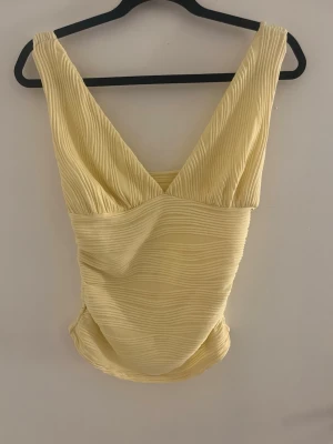 Ljusgul ribbad linnetopp - Snygg ljusgul linnetopp med ribbad struktur och breda axelband. Toppen har en djup v-ringning både fram och bak, vilket ger en trendig och fräsch look. Perfekt att matcha med jeans eller kjol för en cool sommarstil.