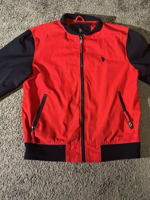 Röd jacka från U.S Polo Assn - Snygg röd jacka från U.S Polo Ass  med svarta ärmar och detaljer. Jackan har dragkedja framtill, två snedställda fickor med dragkedja och ribbade muddar. Klassisk broderad logga på bröstet. Perfekt för dig som vill sticka ut med stil.