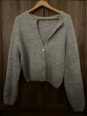 Grå stickad kofta  - Mysig grå stickad kofta med långa ärmar och ribbade muddar. Koftan har en avslappnad passform och stängs med pärlemorskimrande knappar framtill. Perfekt att slänga över en t-shirt för en chill look.