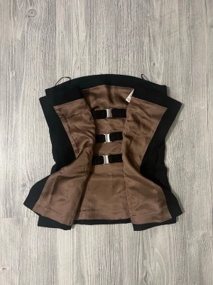 Svart axelbandslös korsett - Stilren svart korsett med axelbandslös design och bruna innerdetaljer. Insidan har tre svarta spännen för att hålla korsetten på plats. Utsidan är slät och figurnära, perfekt för att skapa en snygg siluett. Materialet känns mjukt och har en lätt glans. Köpt för ny år men hittade något annat så nu är den inte till någon användning. Köpte för 659kr säljer för 350kr