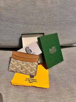 Beige och brun korthållare från G - Snygg korthållare från G@yard i beige och brunt med det klassiska G-mönstret. Tillverkad i canvas och läder, har flera fack för kort och är perfekt för dig som vill ha en stilren och exklusiv accessoar. Kommer med originalask, duk och G kort.