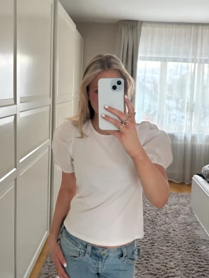 Vit topp med puffärmar - Säljer en stilren vit topp med puffiga korta ärmar och rund halsringning. T-shirten har en feminin och trendig siluett som passar perfekt till jeans eller kjol. Perfekt för dig som gillar en enkel men snygg look.