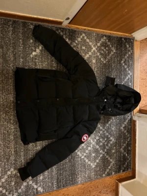 Svart dunjacka från Canada Goose - Säljer en svart dunjacka från Canada Goose med huva och klassisk patch på ärmen. Jackan har dragkedja och knappar framtill, flera fickor och en riktigt puffig look. Perfekt för kalla vinterdagar och riktigt varm.