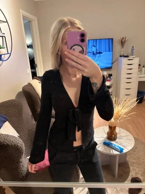 Svart knytblus med djup urringning - Trendig svart blus med lång ärm och djup urringning framtill. Blusen har en snygg knytning vid bröstkorgen som ger en cool och modern look. Perfekt att styla med jeans eller kjol för en chic outfit.