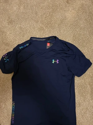 Blå tränings-tshirt från Under Armour - Snygg mörkblå tränings-tshirt från Under Armour med regnbågsfärgat logotryck på bröstet och textdetaljer på ärmen och sidan. Tillverkad i lätt och ventilerande HeatGear-material som håller dig sval under träningen. Perfekt för löpning eller gym.