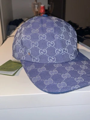 Blå GG-mönstrad keps från HM - Snygg blå keps från HM med det klassiska GG-monogrammet över hela kepsen. Kepsen har böjd skärm och är tillverkad i ett tåligt tygmaterial. Perfekt för dig som vill sticka ut med en exklusiv accessoar.